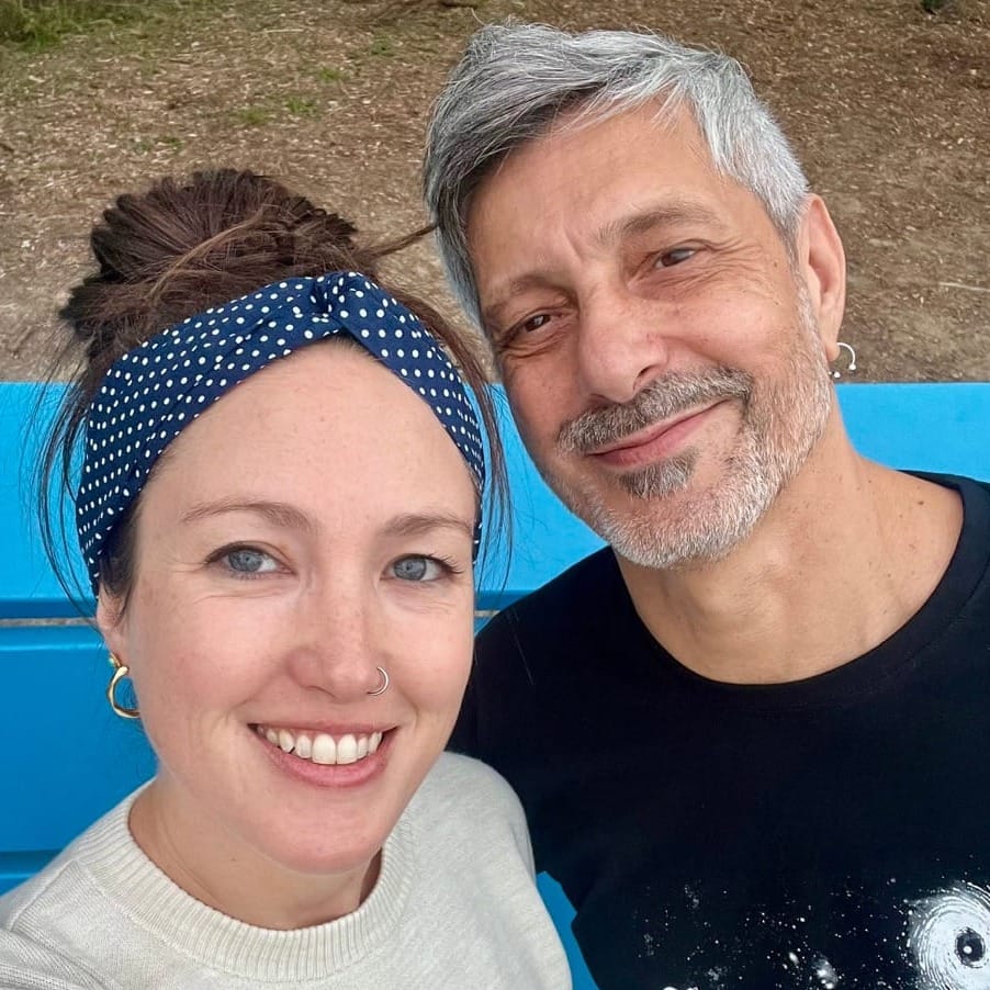 A smiling woman with blue eyes, a cream top, and polkadot headband poses for a selfie with a smiling, grey-haired man with a beard and a black t-shirt